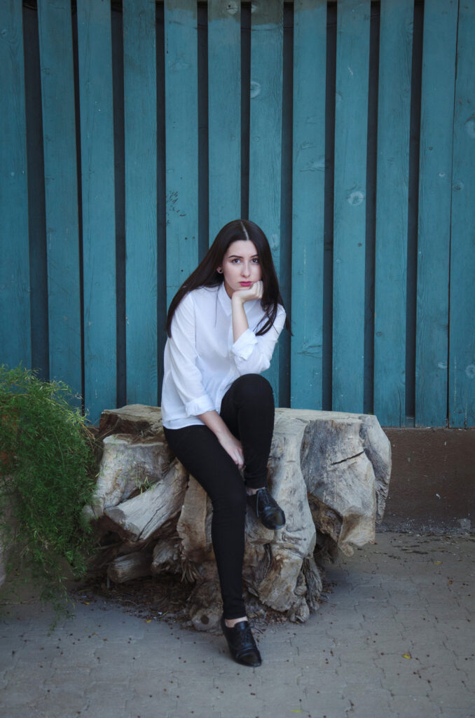 Gabriela Inzunza sits pensively on a weathered wooden stump against a teal wooden backdrop with a green plant to her left.
